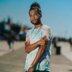A fashionable adult male stands confidently on a sunny boardwalk in New York.