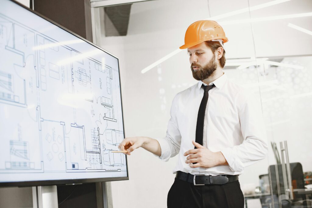 Architect in hard hat reviewing blueprints on a screen, discussing plans in modern office.