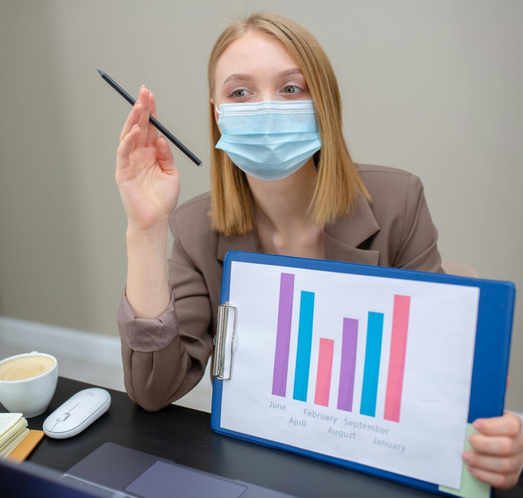Young woman in office wearing face mask, presenting graphs, adapting to new normal work environment.